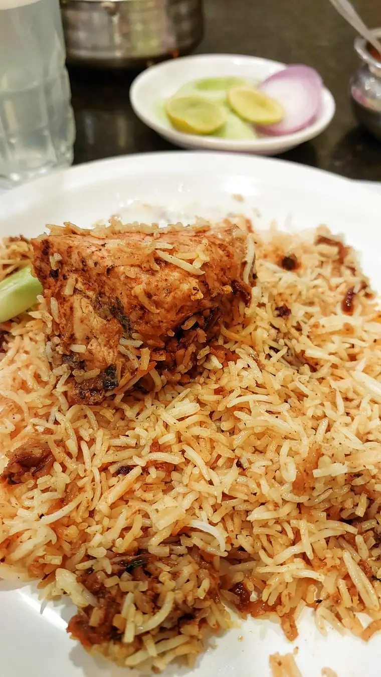 A plate of biryani with a large piece of meat, onions and lime on the side