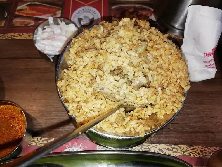 A wide steel vessel of seeraga samba biryani with short, separate grains, served with onion raita